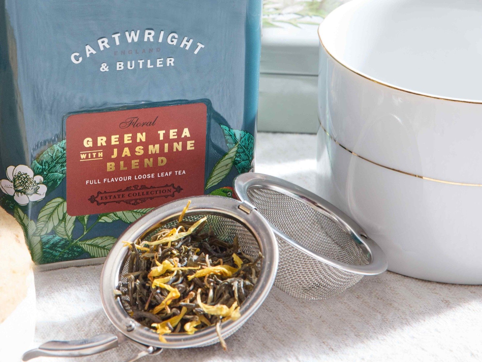 Green Tea with Jasmind Blend Tea in a tea strainer next to a cartwright & butler tin and teacup