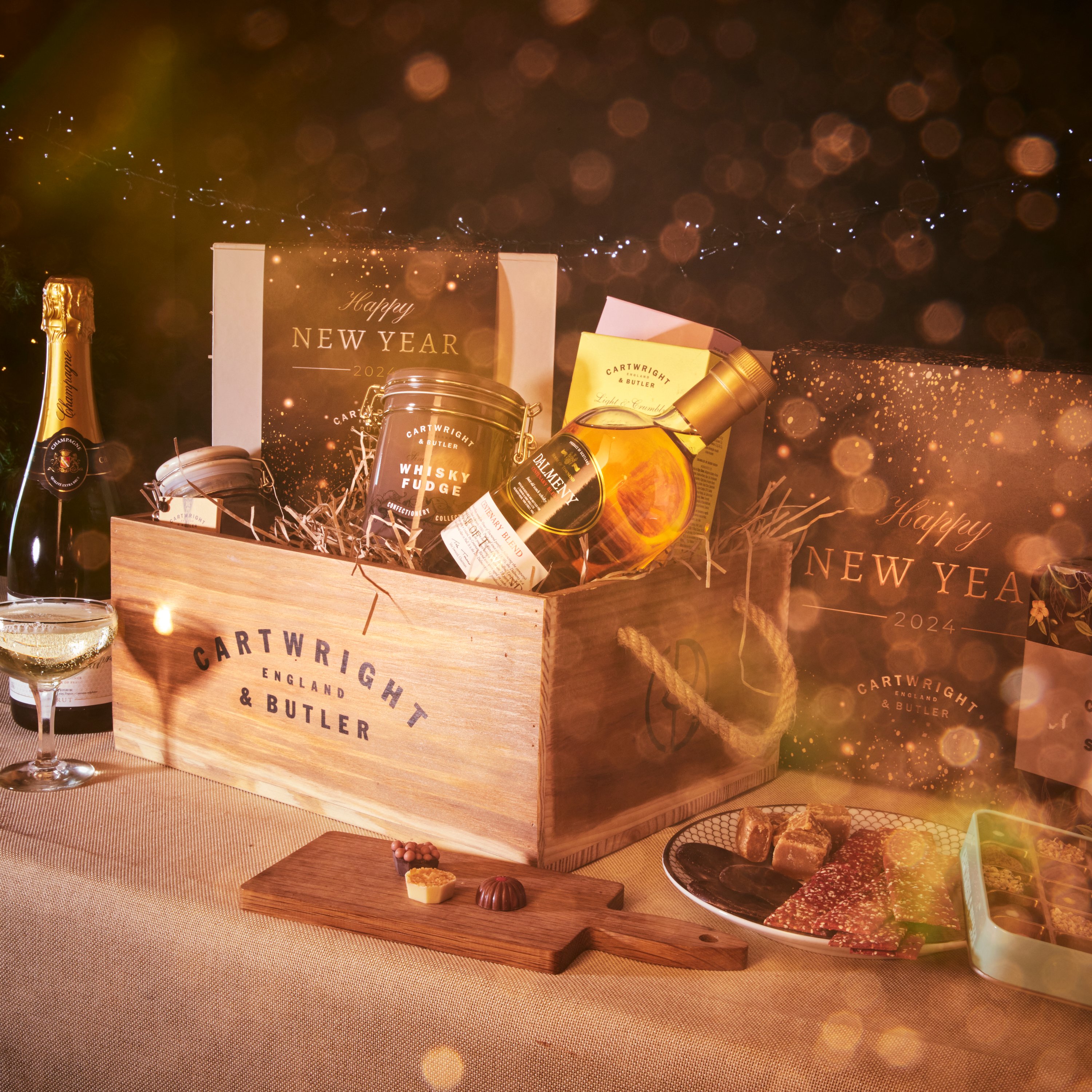 A festive New Year's Eve hamper filled with gourmet treats and a bottle of champagne on a table decorated with lights.