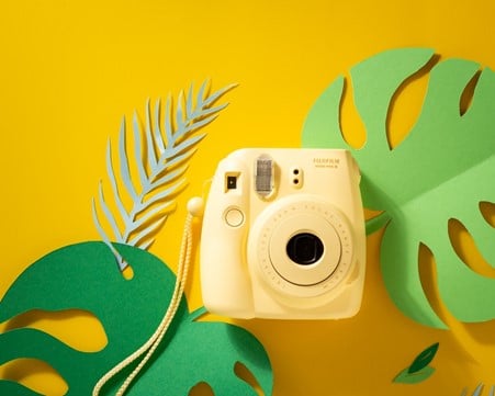Cream polaroid camera set amongst yellow background, with blue and green paper leaf stencils