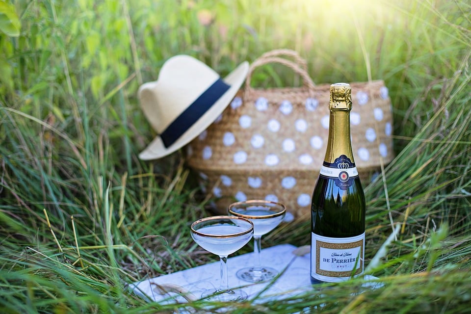 Spotted wicker picnic basket in a field with two glasses and bottle of champagne